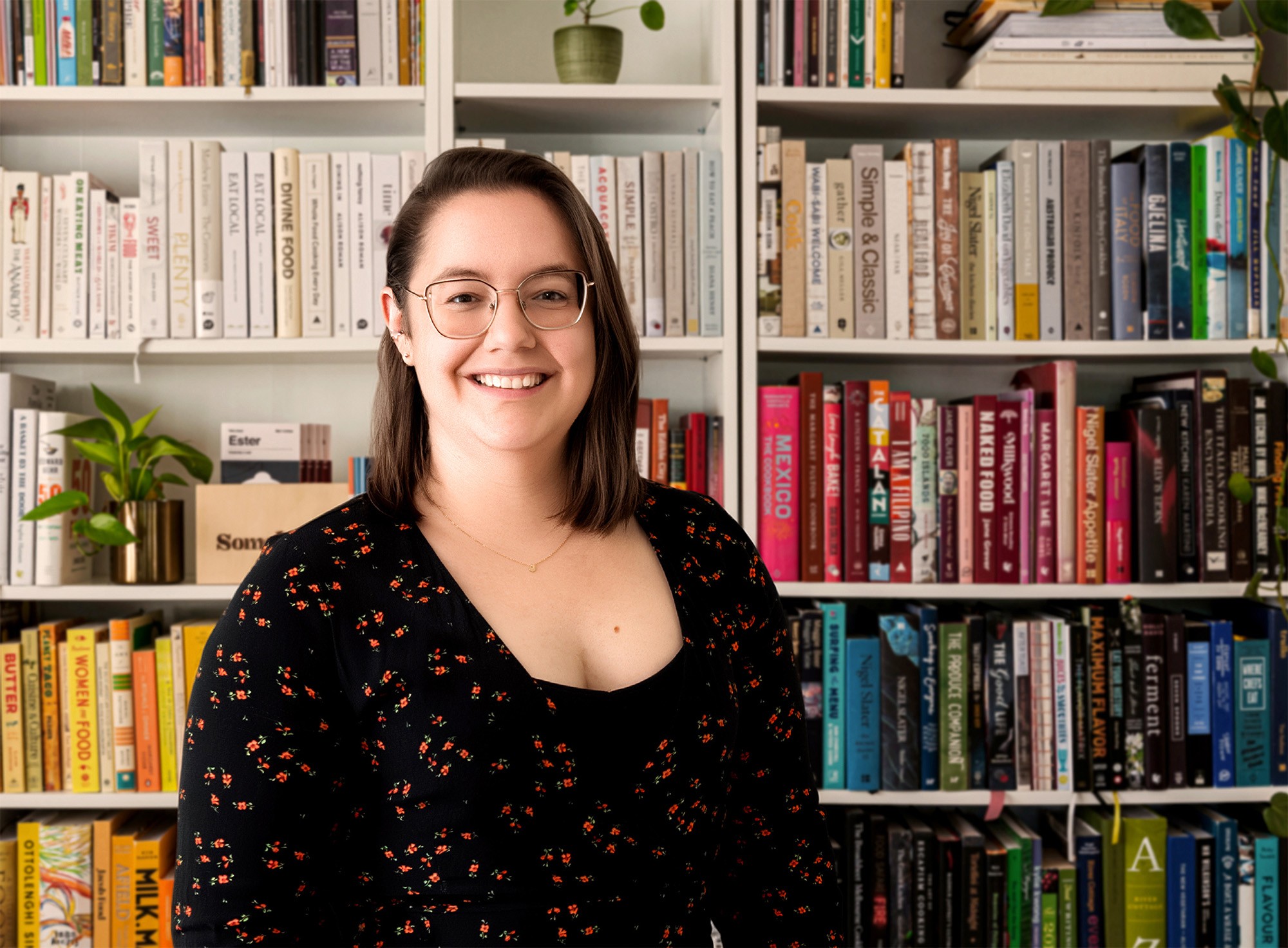 portrait of a smiling woman with brown hair, wearing glasses and a black long-sleeved dress, standing in front of a white bookshelf